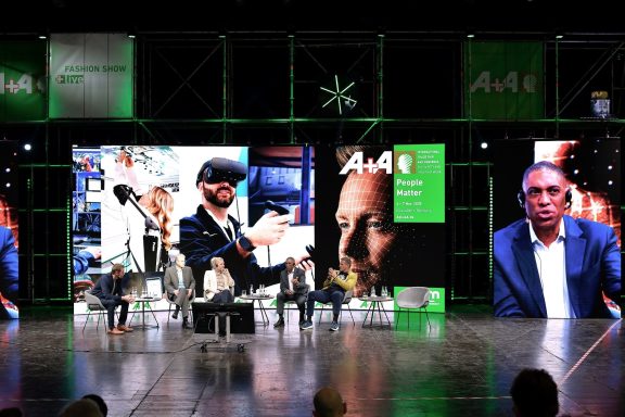 Bühne mit Moderatoren, Präsentationen und großen Bildschirmen bei einer Veranstaltung.